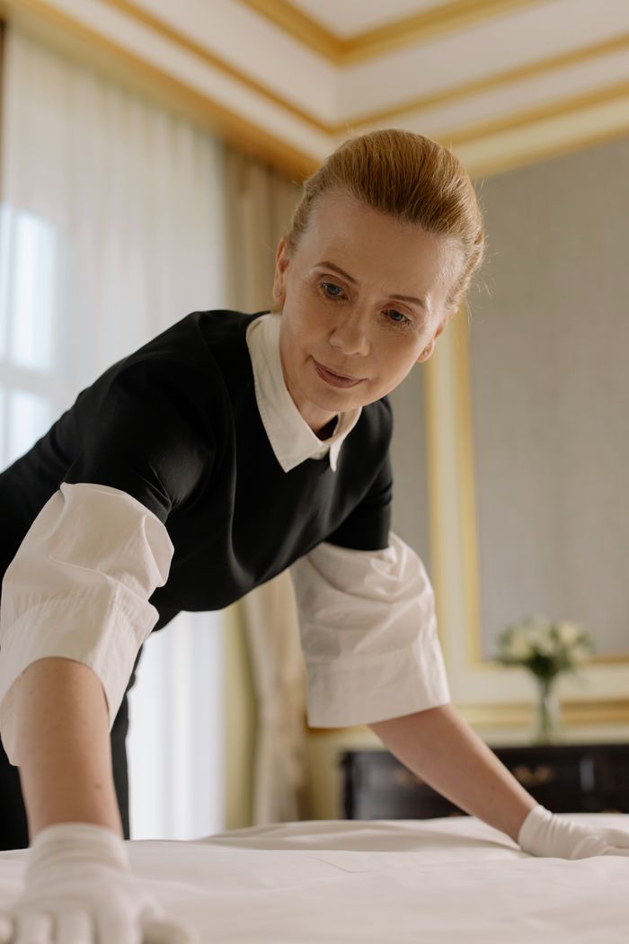 Une femme de chambre en uniforme dispose avec adresse le linge de lit dans une chambre d’hôtel luxueuse.